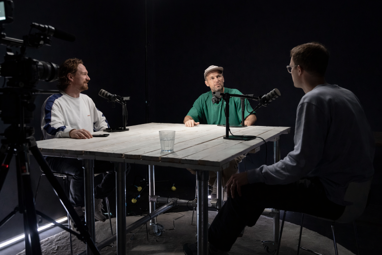 Twee podcasters nemen een aflevering op in een professionele podcaststudio met microfoons en camera-opstelling.